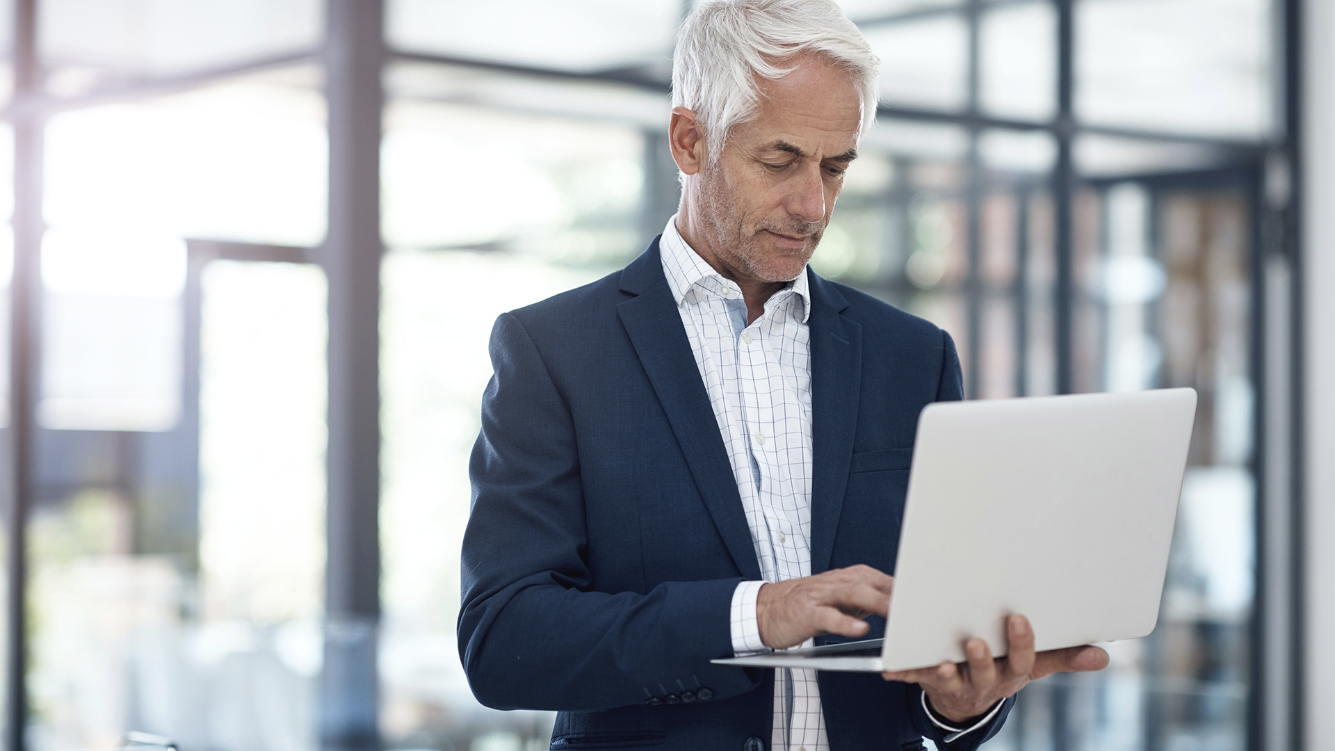 An older man with gray hair in a dark suit and white shirt stands in a bright office environment, focused on working on a laptop. Large windows and a modern interior are visible in the background.