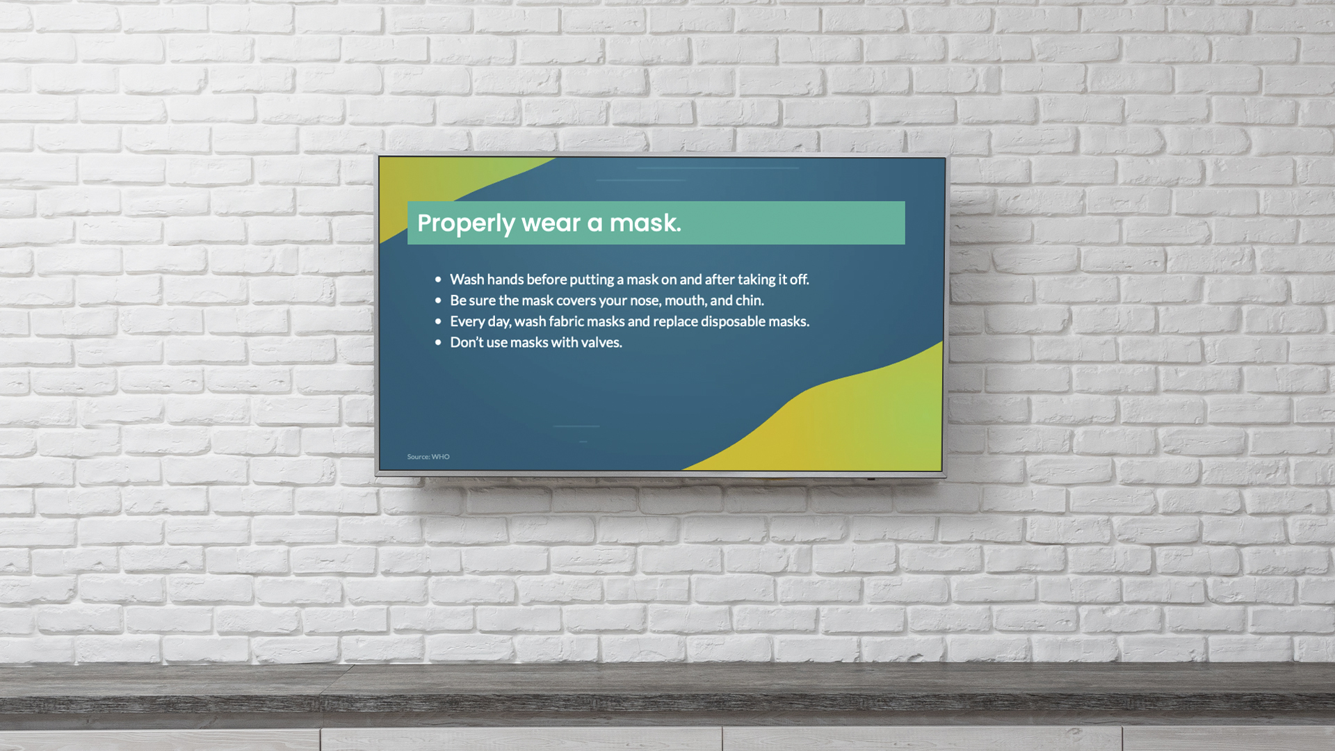 A TV mounted on a white brick wall displays instructions on properly wearing a mask. The text includes tips such as washing hands, covering the nose and mouth, and avoiding masks with valves. The background features a green and yellow design.