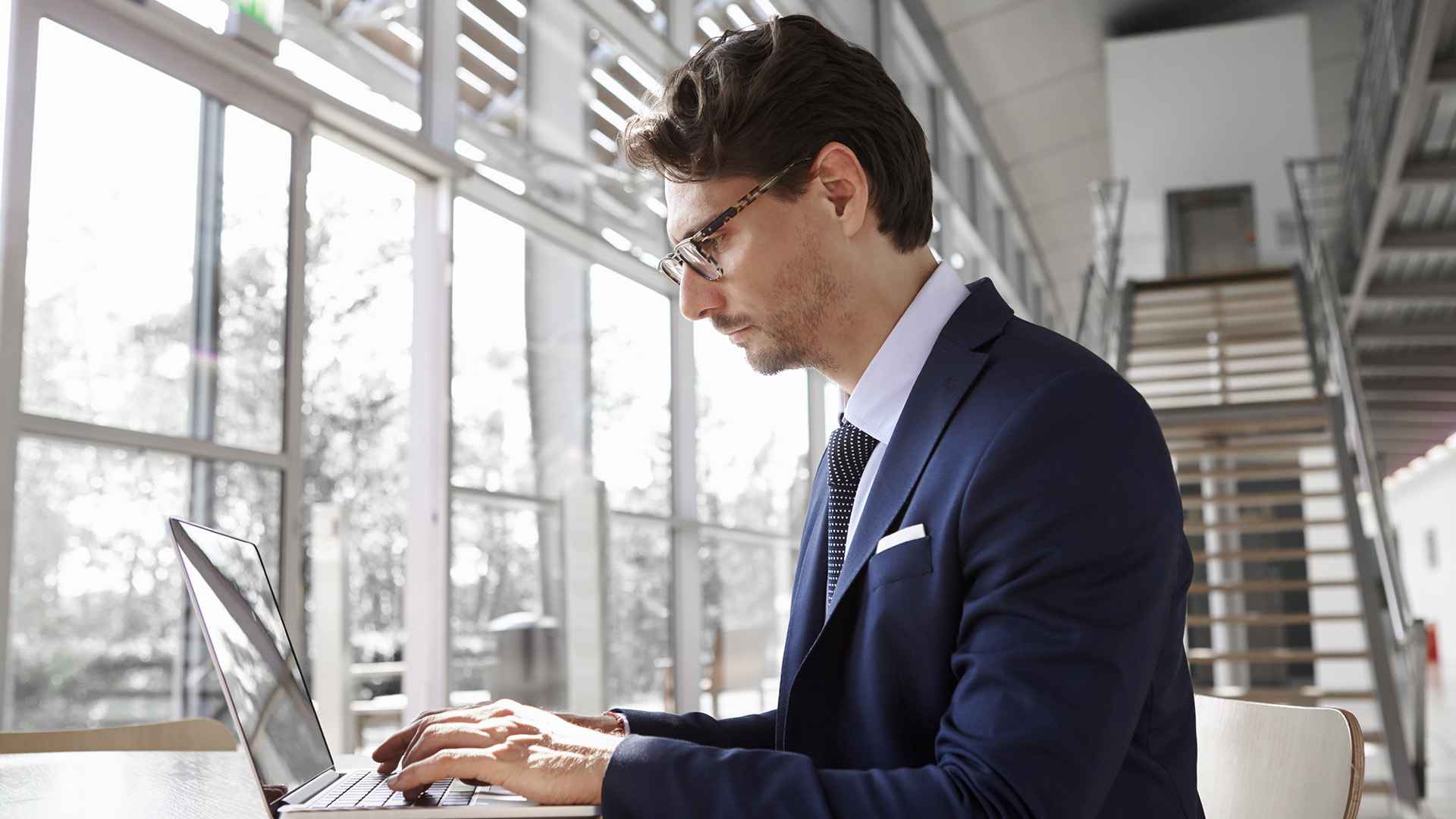 A professionally dressed man in a navy suit working on a laptop in a bright, modern office.
