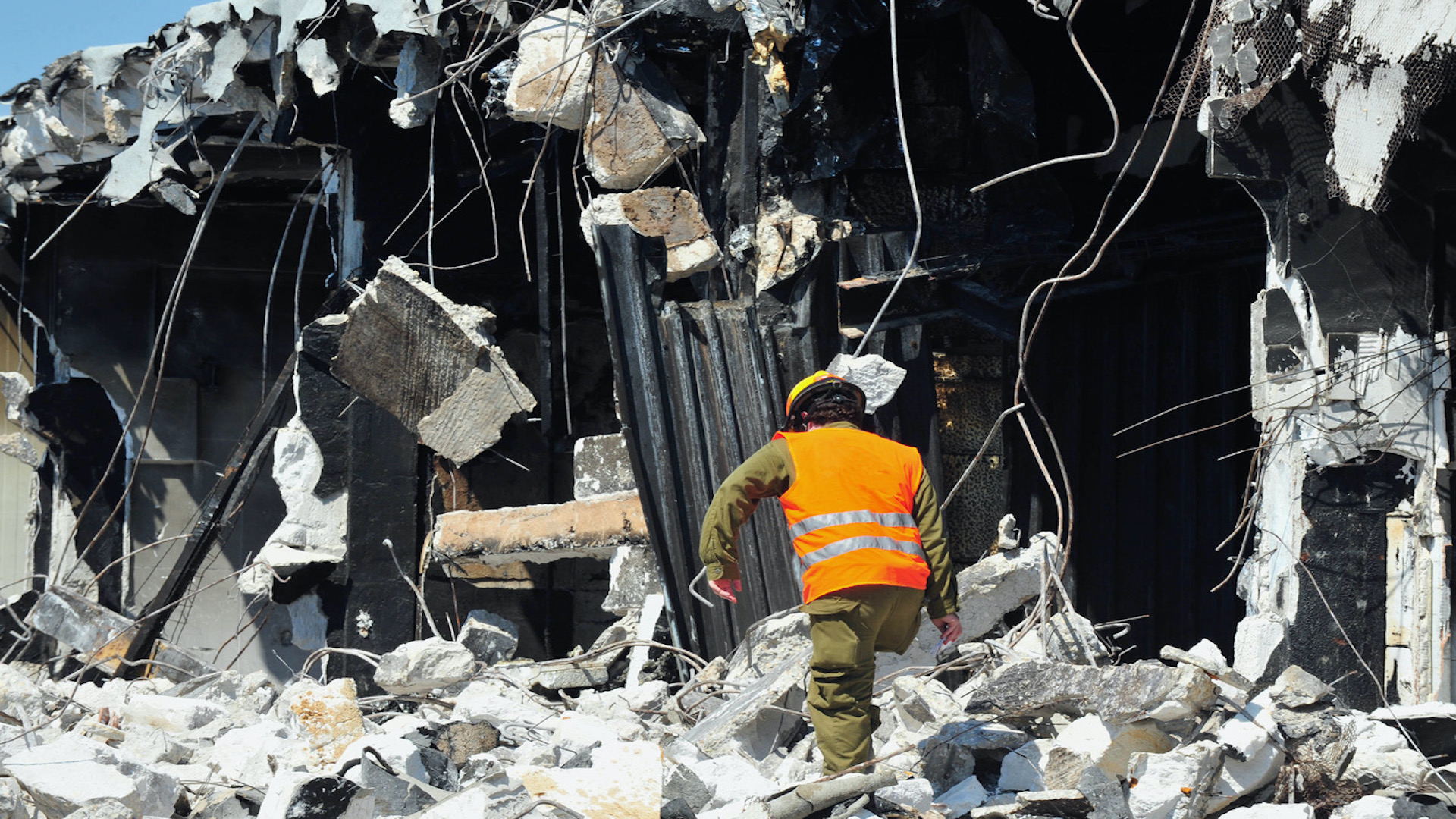 A worker in a high-visibility orange vest and helmet navigates through the rubble of a severely damaged building, with debris and twisted metal scattered around. The scene suggests a post-disaster environment.