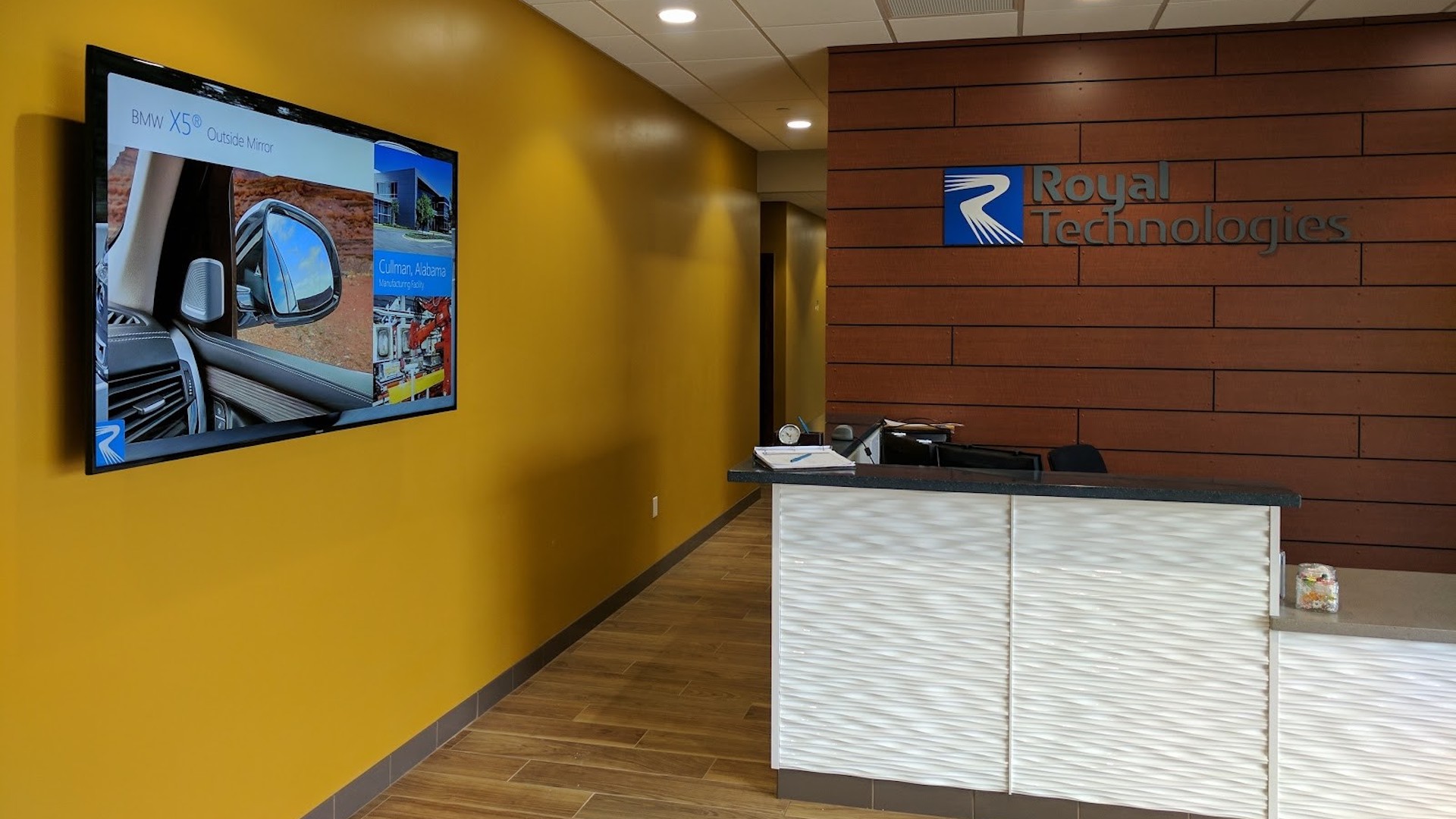 Reception area of Royal Technologies with a modern design. A wall-mounted screen displays various images and text. The front desk features a textured white surface, and the company logo is on the wooden wall. Warm lighting creates an inviting atmosphere.