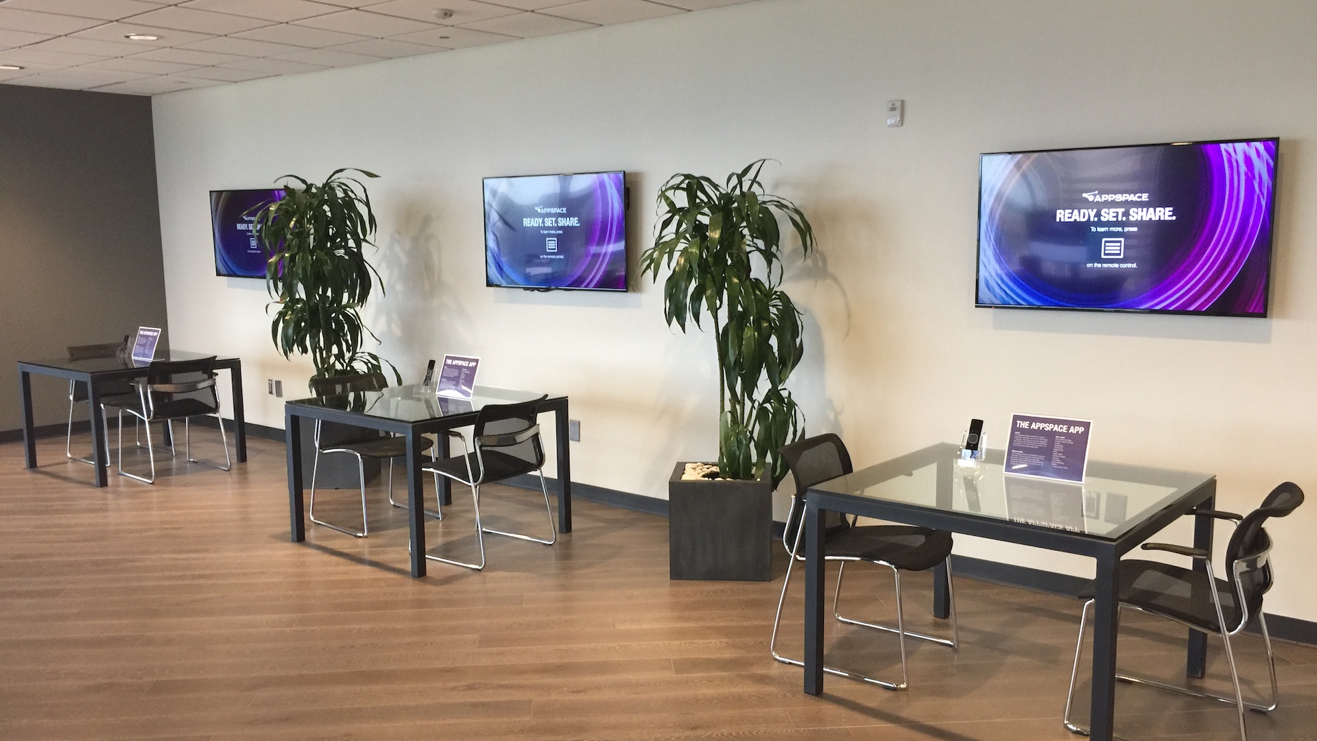 A modern waiting area with four glass-top tables and chairs against a wall. Three flat-screen TVs display READY. SET. SHARE. Plant decorations are in between. Wood flooring and overhead lighting complete the sleek look.