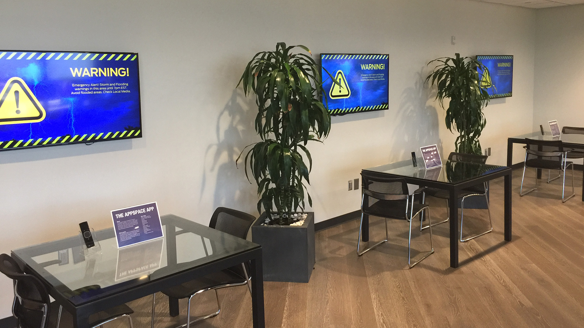 A lounge area with glass tables, chairs, and large digital screens displaying a storm and flooding warning message.