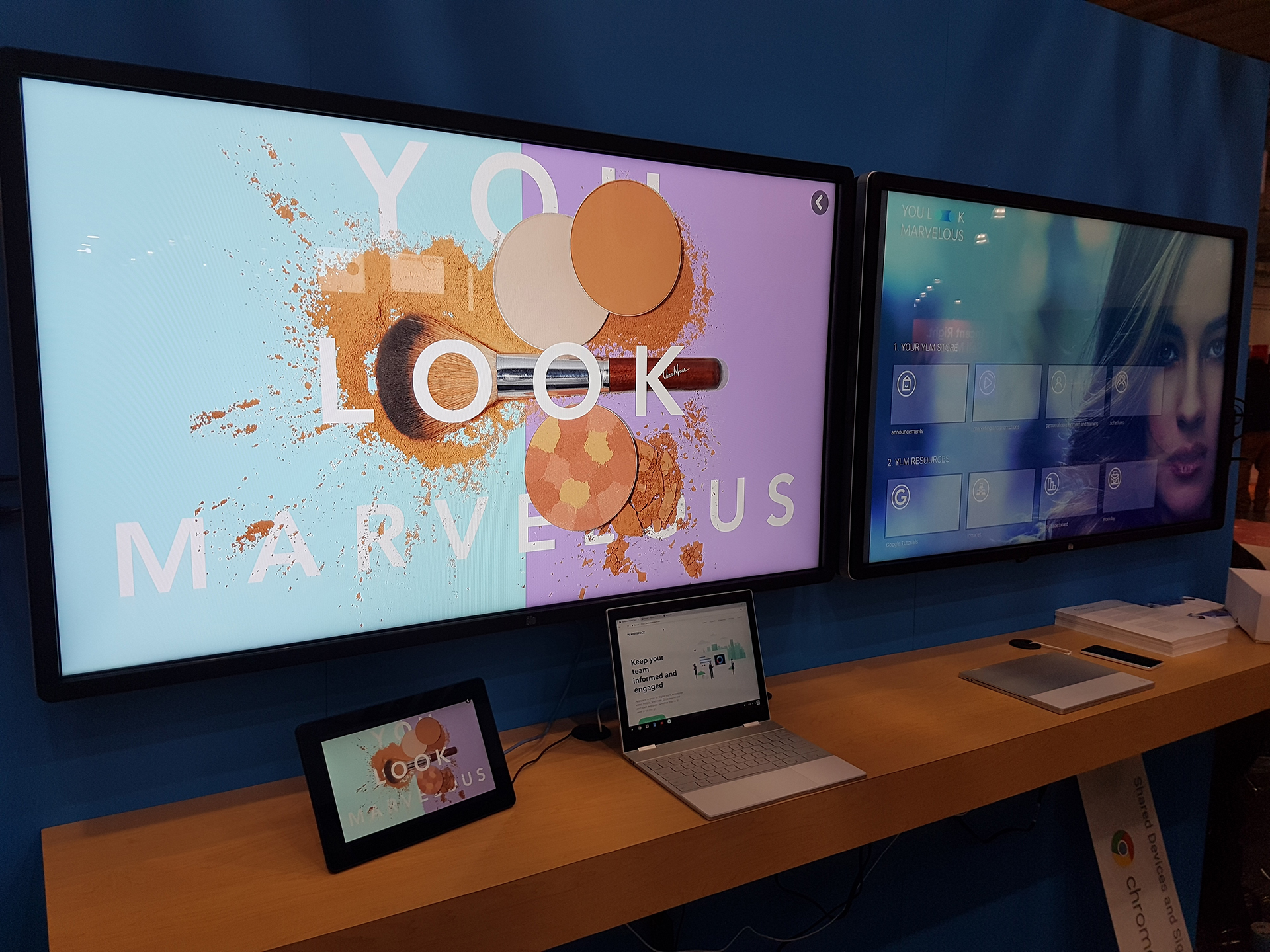 Two large displays show interactive content. The left screen features makeup items with the text YOU LOOK MARVELOUS. Below, a tablet and laptop display the same image. The right screen shows an interface with multiple icons. A wooden shelf holds the devices.