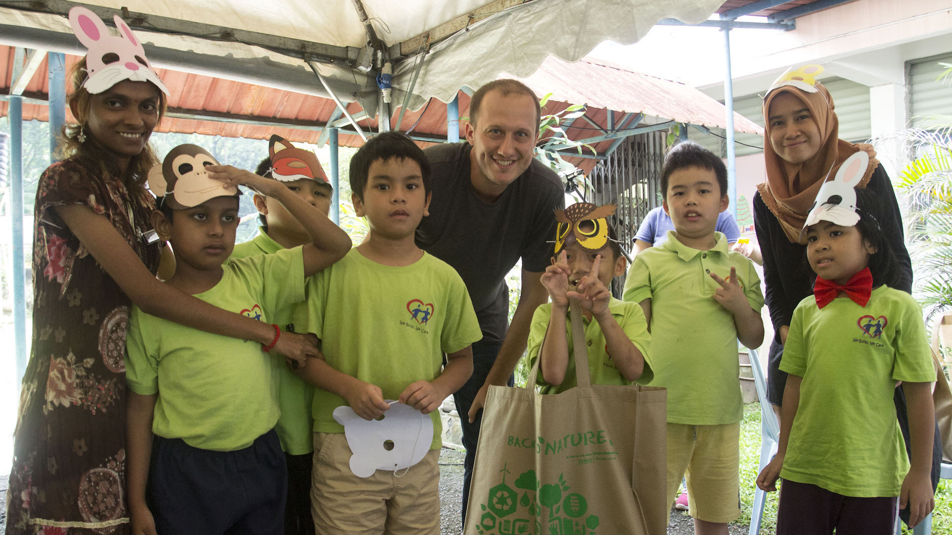 A group of children in green shirts and animal masks pose with two adults under a canopy. The adults are smiling, and one child holds a bag. The setting appears to be an outdoor event.