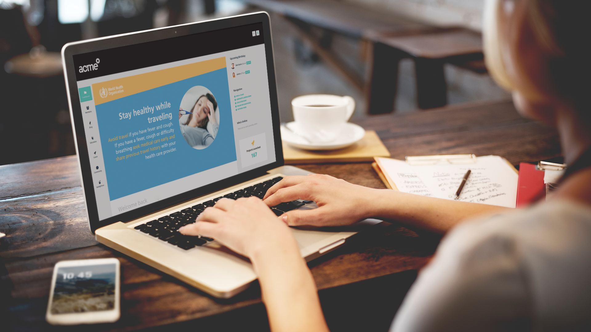 A person is using a laptop at a cafe table. The screen displays a website with a health-themed slide titled Stay healthy while traveling. A cup of coffee, a notepad, and a smartphone showing 10:45 are on the table.