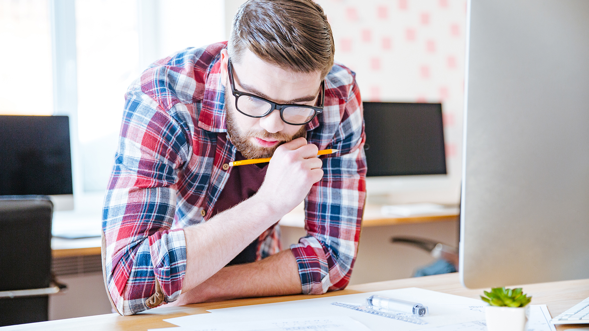 A man with glasses and a plaid shirt concentrates on work at a desk, holding a pencil to his mouth. He is leaning over papers with a focused expression. Computer monitors and a small potted plant are visible in the background.