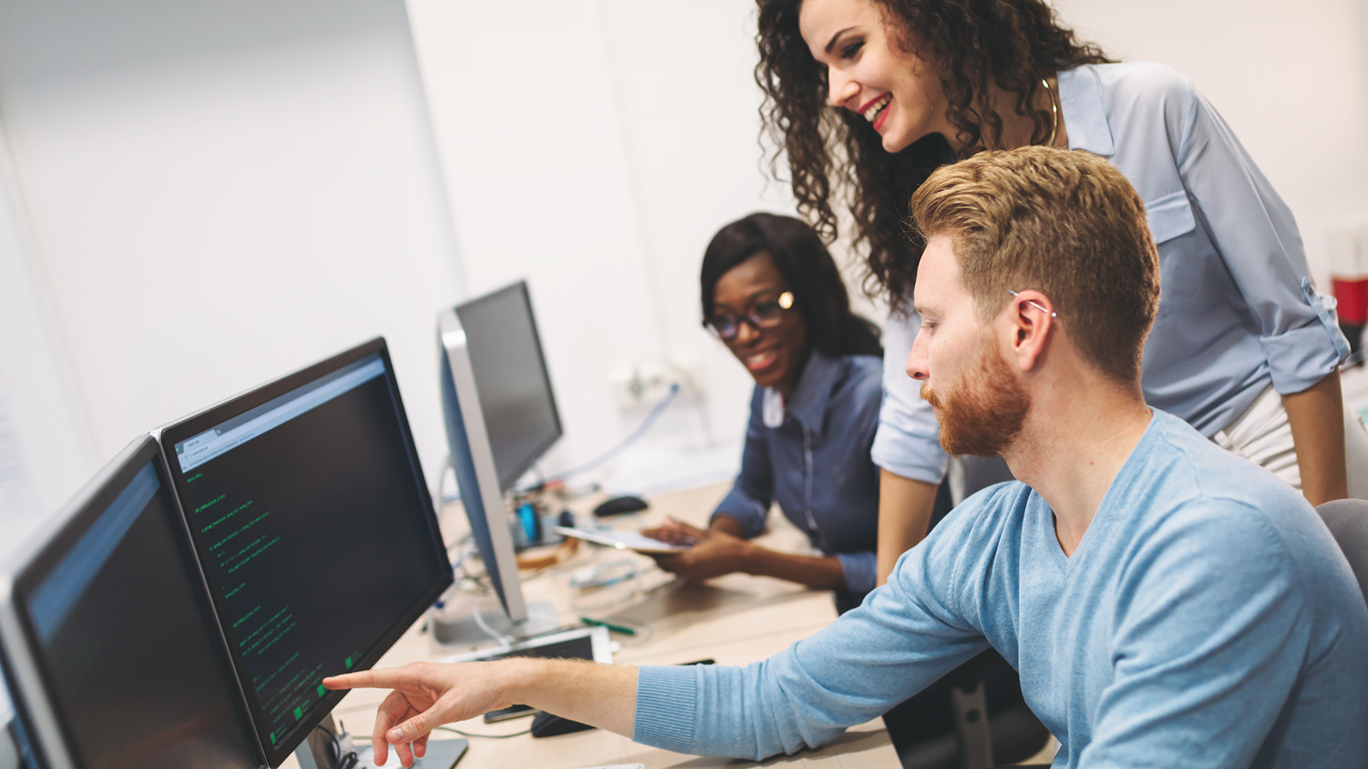 Three colleagues collaborating in an office, analyzing code on dual computer monitors.