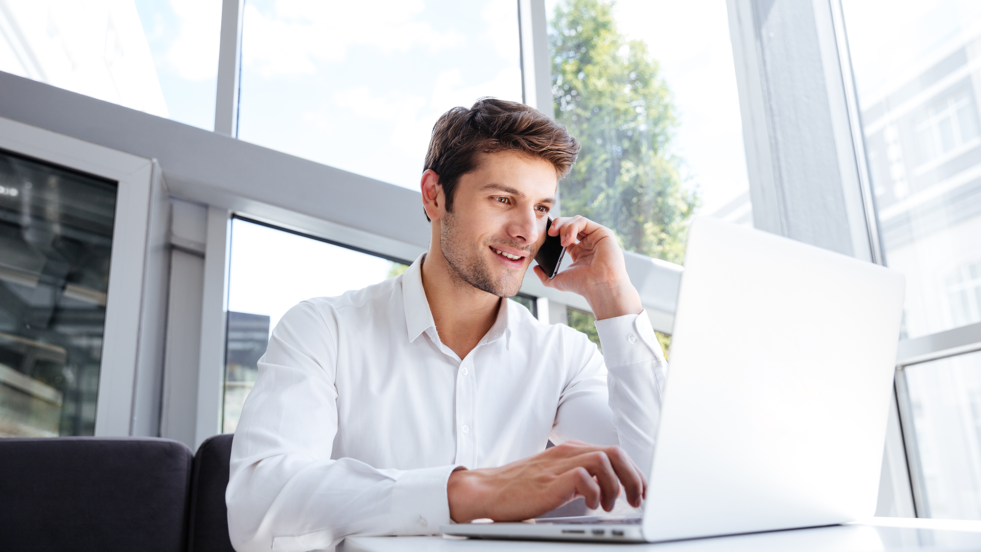 A man in a white shirt is smiling and talking on a smartphone while working on a laptop. He is seated in a modern, bright office with large windows showing a view of trees outside.