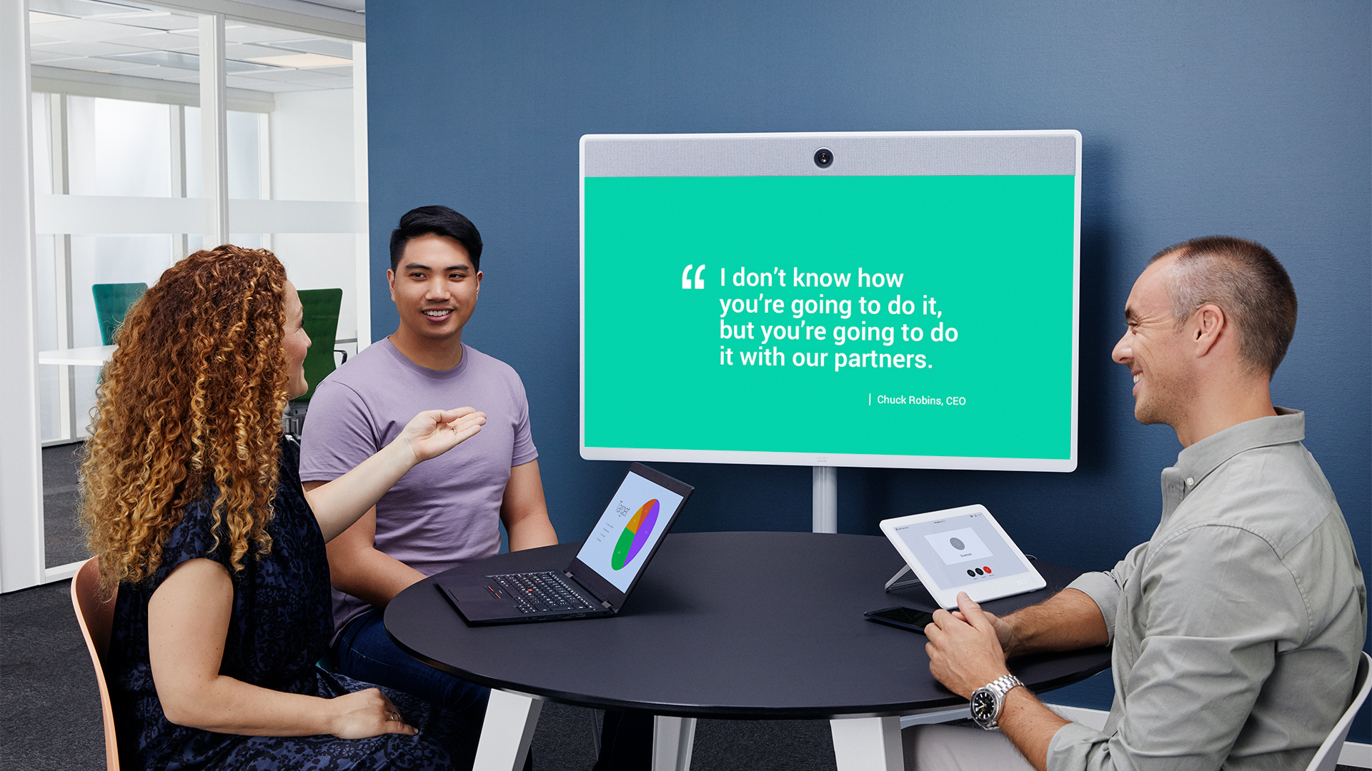 Three people sit at a round table in an office. They have laptops and tablets. A large screen shows the quote, I dont know how youre going to do it, but youre going to do it with our partners.