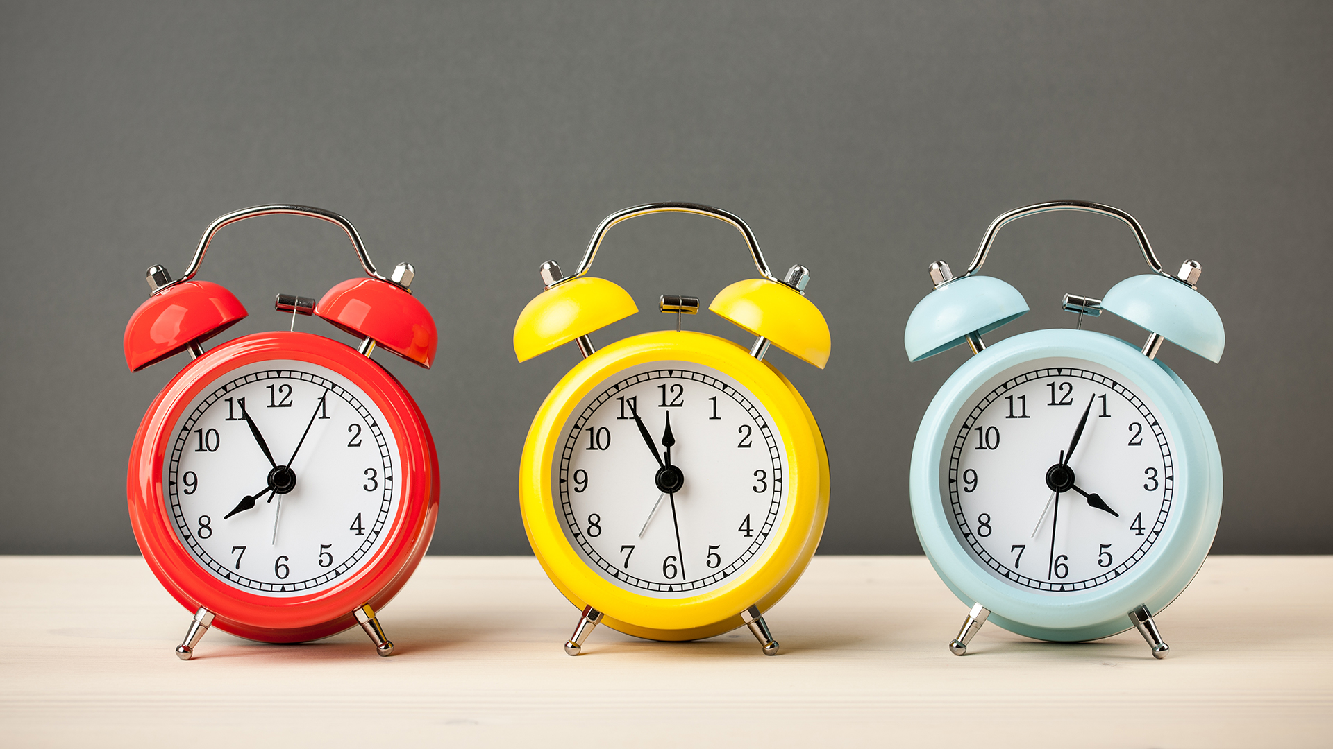 Three colorful vintage alarm clocks sit in a row on a light wooden surface against a gray background. The clocks are red, yellow, and blue, each showing the same time: 10:10.