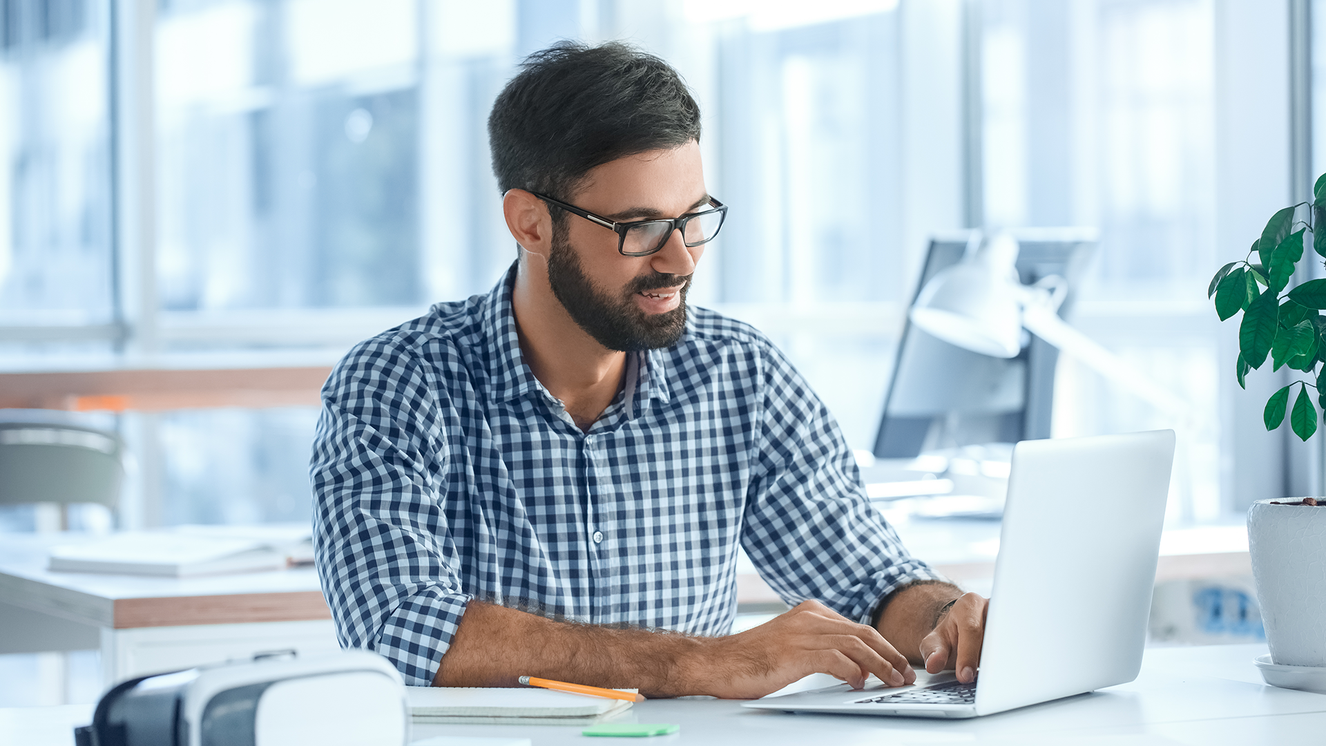 Man with a beard and glasses seated at a desk, typing on a laptop. Hes wearing a checkered shirt and is in a bright office environment with large windows. Nearby are office items, including a plant and a virtual reality headset.