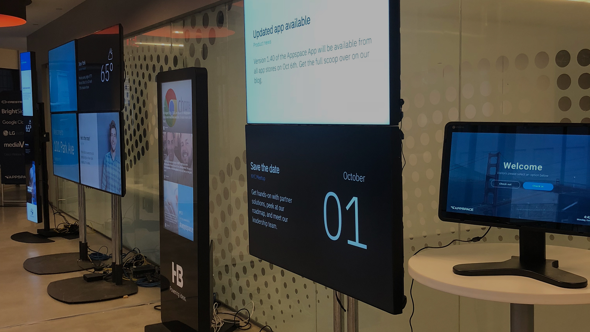 Multiple digital displays in a modern setting show various messages, including a welcome screen, an announcement for an updated app, and a save-the-date for October 1. Cables are visible on the floor, and a reflective glass wall is in the background.