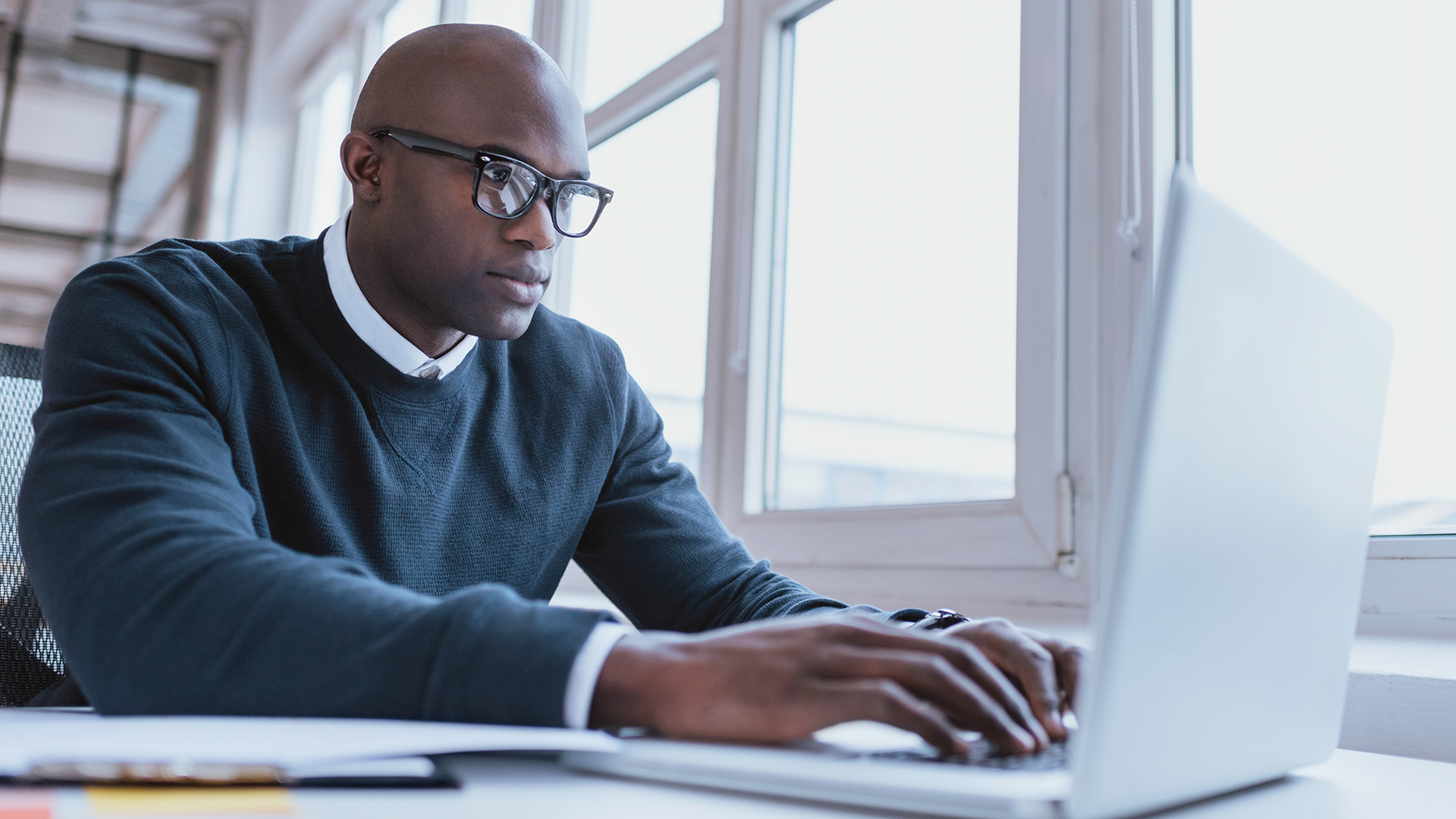 A person wearing glasses and a black sweater works intently on a laptop at a desk. The setting is bright with large windows in the background. Papers and other office items are visible on the desk.