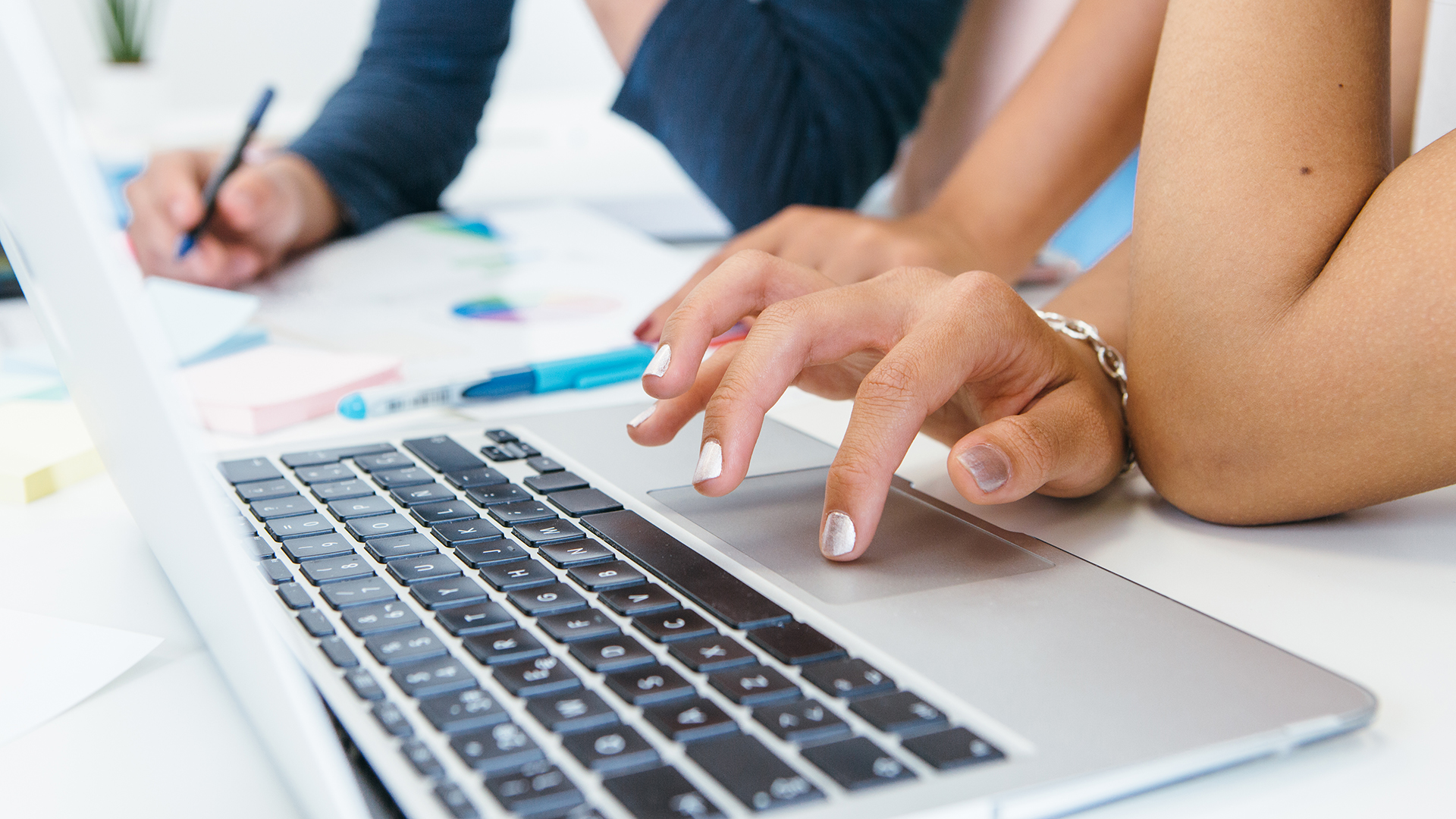Close-up of two people working at a desk. One person types on a laptop while another writes in a notebook. Pens and a notebook are visible on the table. The scene suggests a collaborative or study environment.