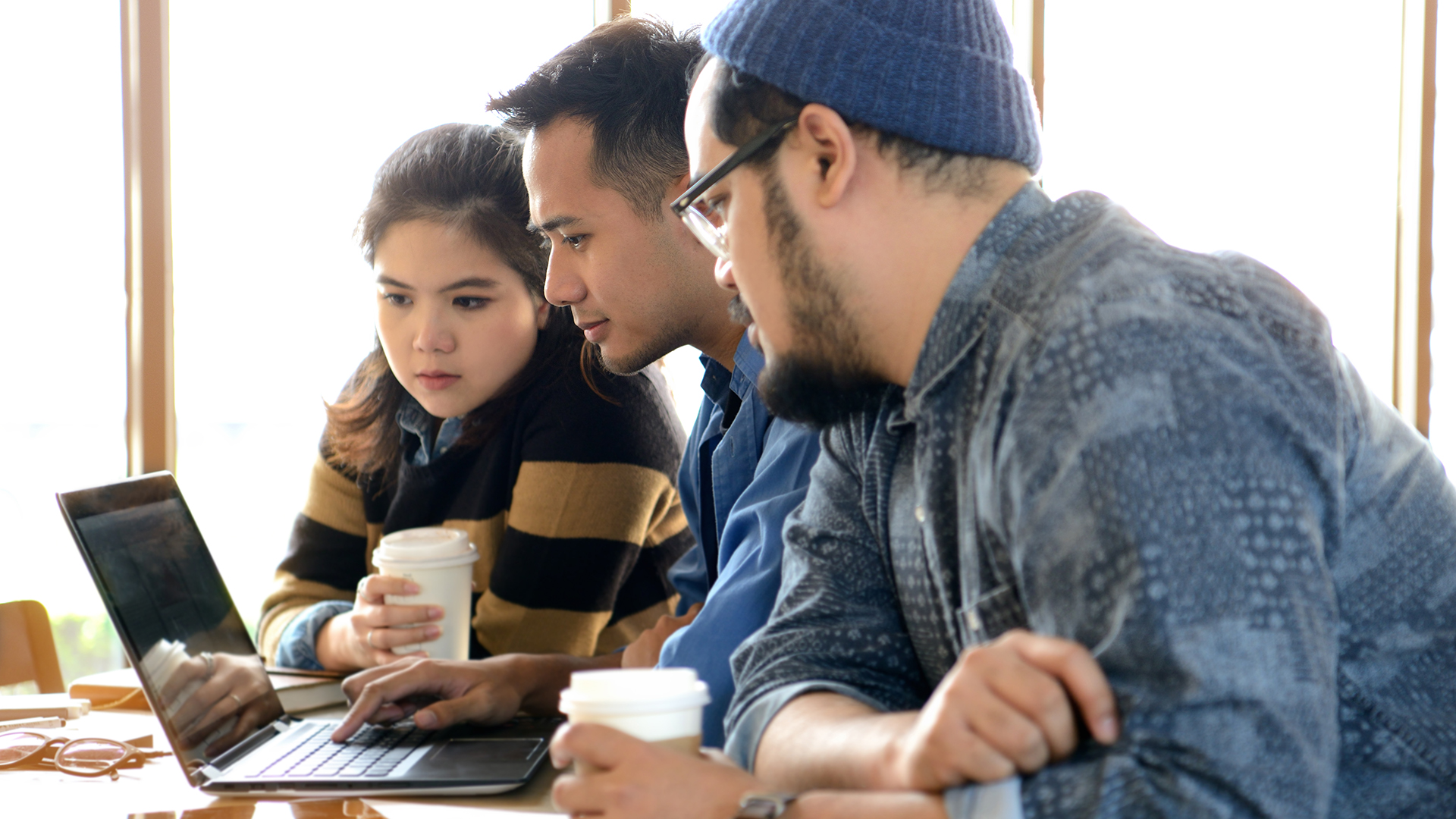 Three people sit at a table in front of a laptop, collaborating. Two men, one wearing glasses and a beanie, and one woman, all focus on the screen. Each holds a coffee cup, with natural light coming in through large windows.
