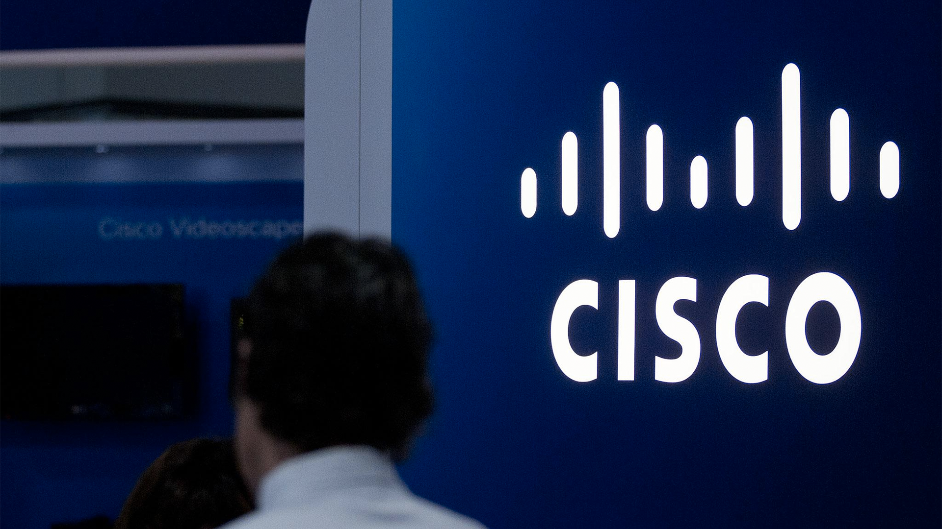 The image shows a blue wall with the Cisco logo in white. A silhouetted person is in the foreground, partially visible. The logo consists of the word CISCO with a vertical line design above it.