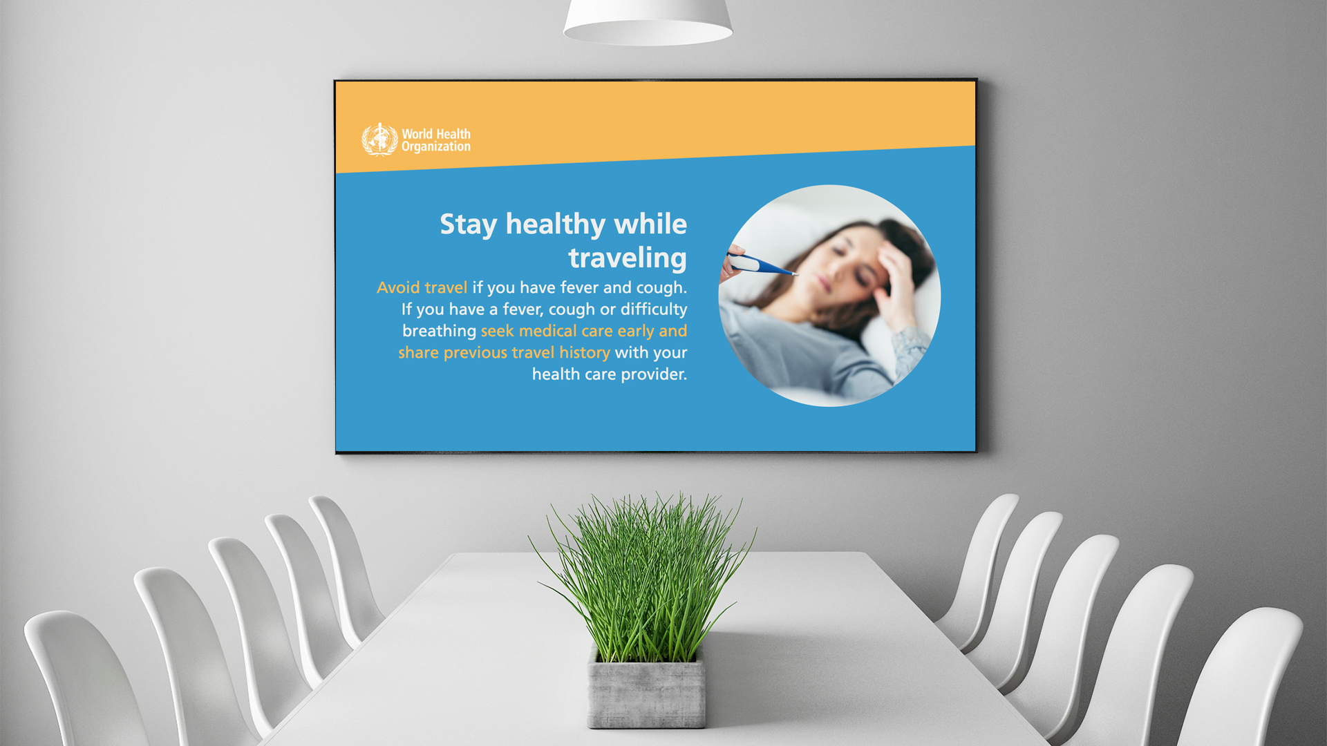 A conference room with a rectangular table and chairs. A screen displays a World Health Organization poster: Stay healthy while traveling with advice to avoid travel if you have a fever or cough and to seek medical care early.