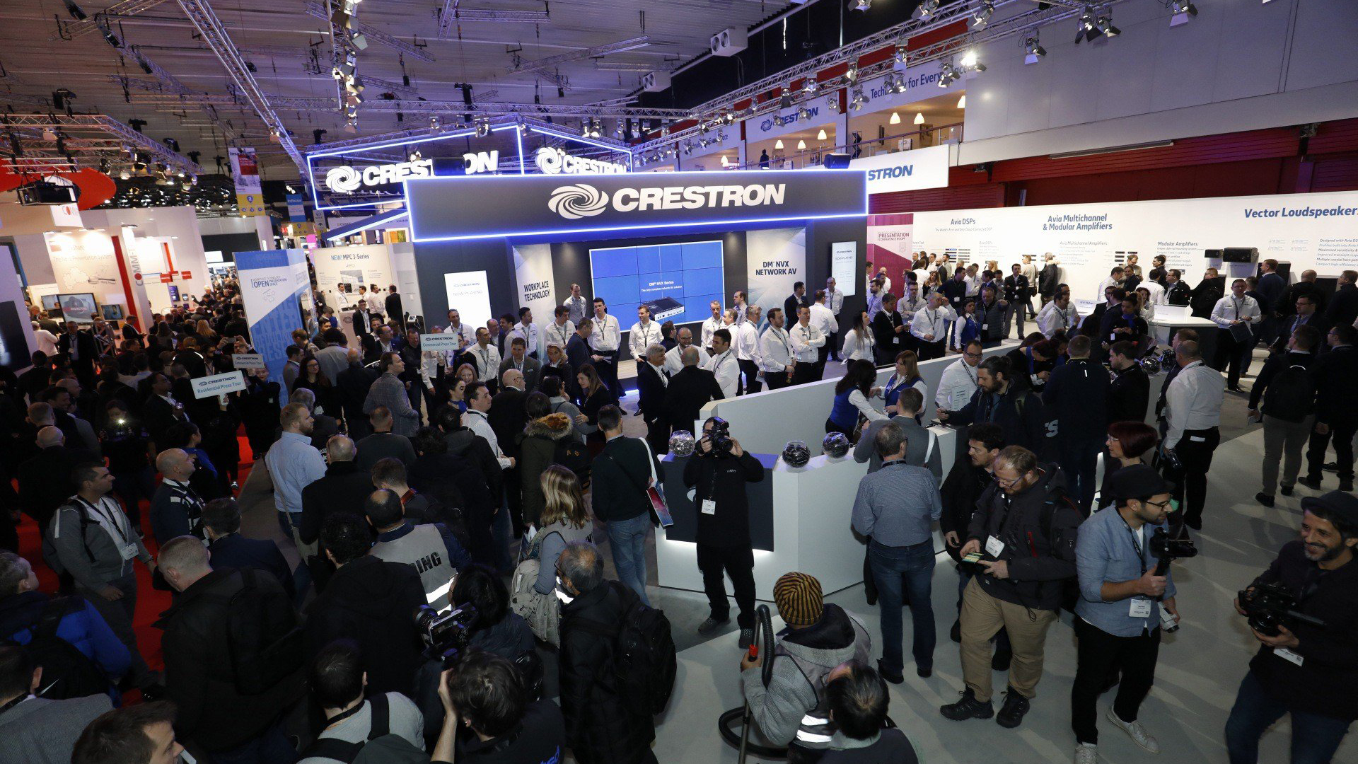 A large crowd at a technology exhibition surrounds the Crestron booth, with many people engaged in conversation and taking photos. Bright overhead lights illuminate the booth and various tech displays are visible in the background.