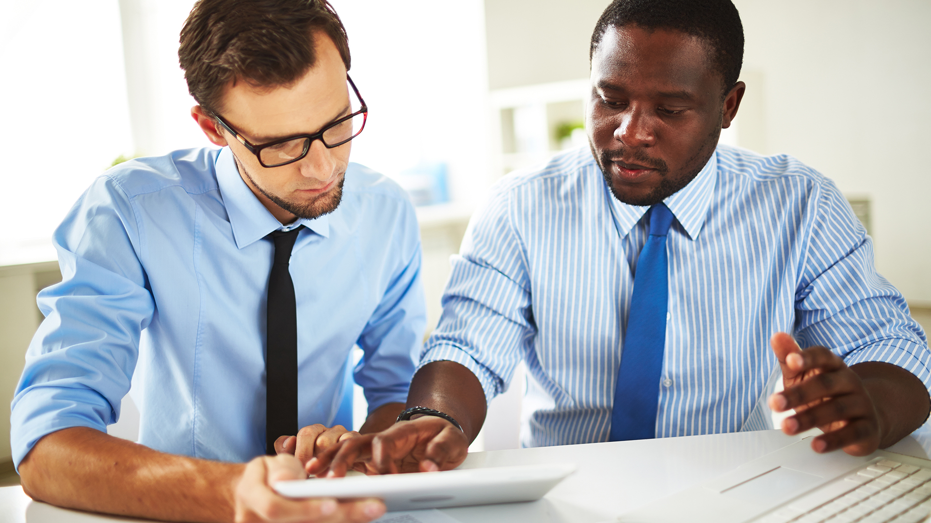 Two men in blue shirts and ties are sitting at a table, focused on a tablet. One man gestures while the other points at the screen, suggesting a collaborative work environment.