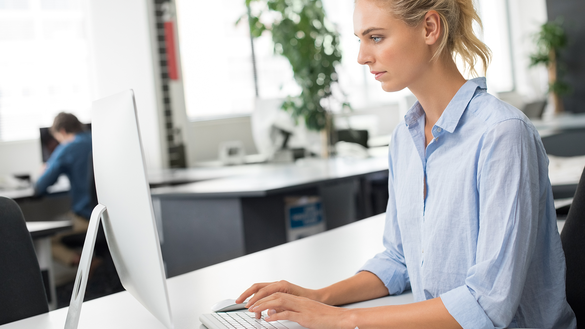 A woman in a light blue shirt is seated at a desk, working on a computer in a modern office setting. Other desks and lush green plants are visible in the background, creating a professional and airy atmosphere.