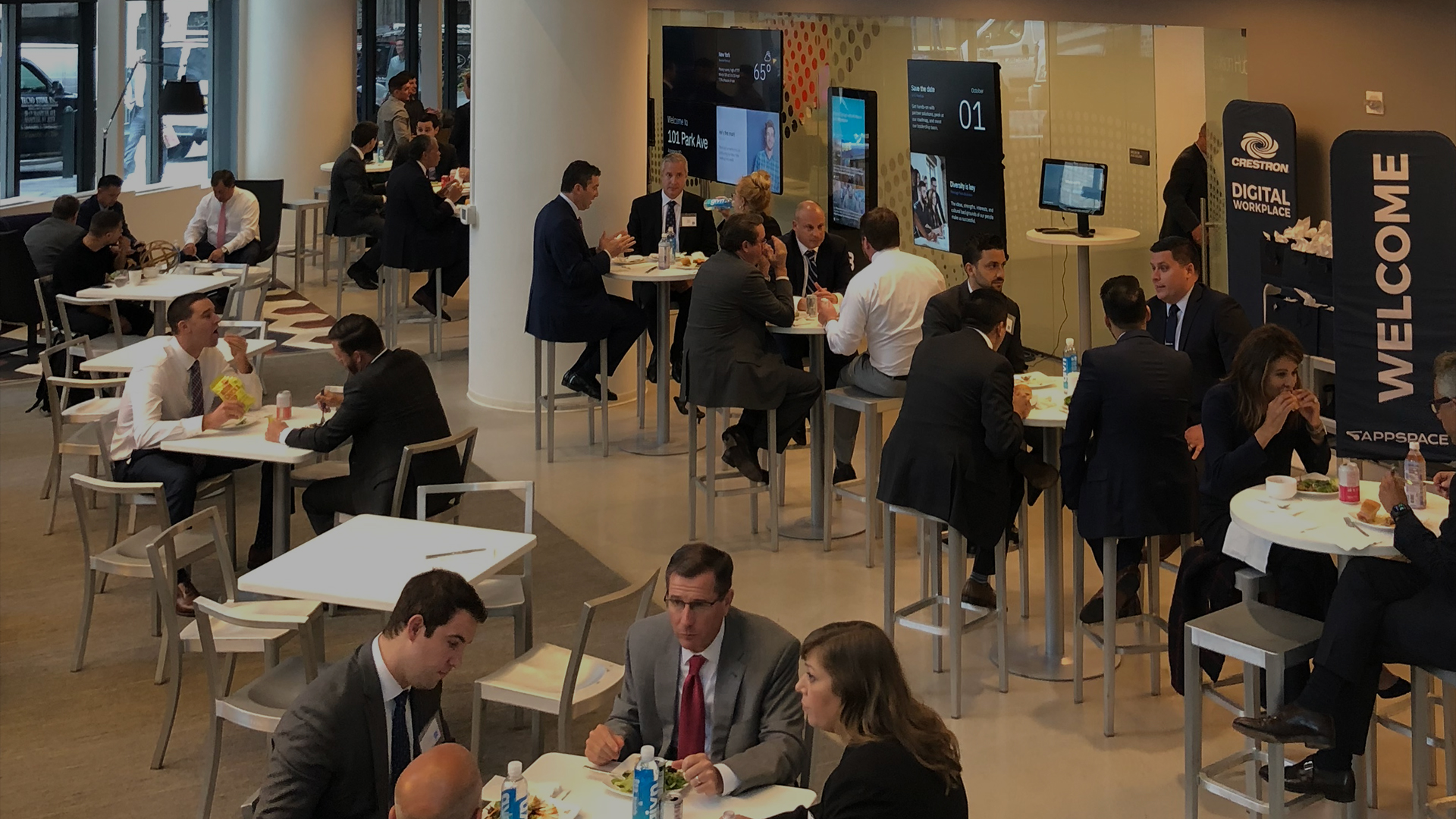 People in business attire sitting and talking around tables in a conference or networking event. Large screens display information on the walls, and a Welcome sign stands in the background.