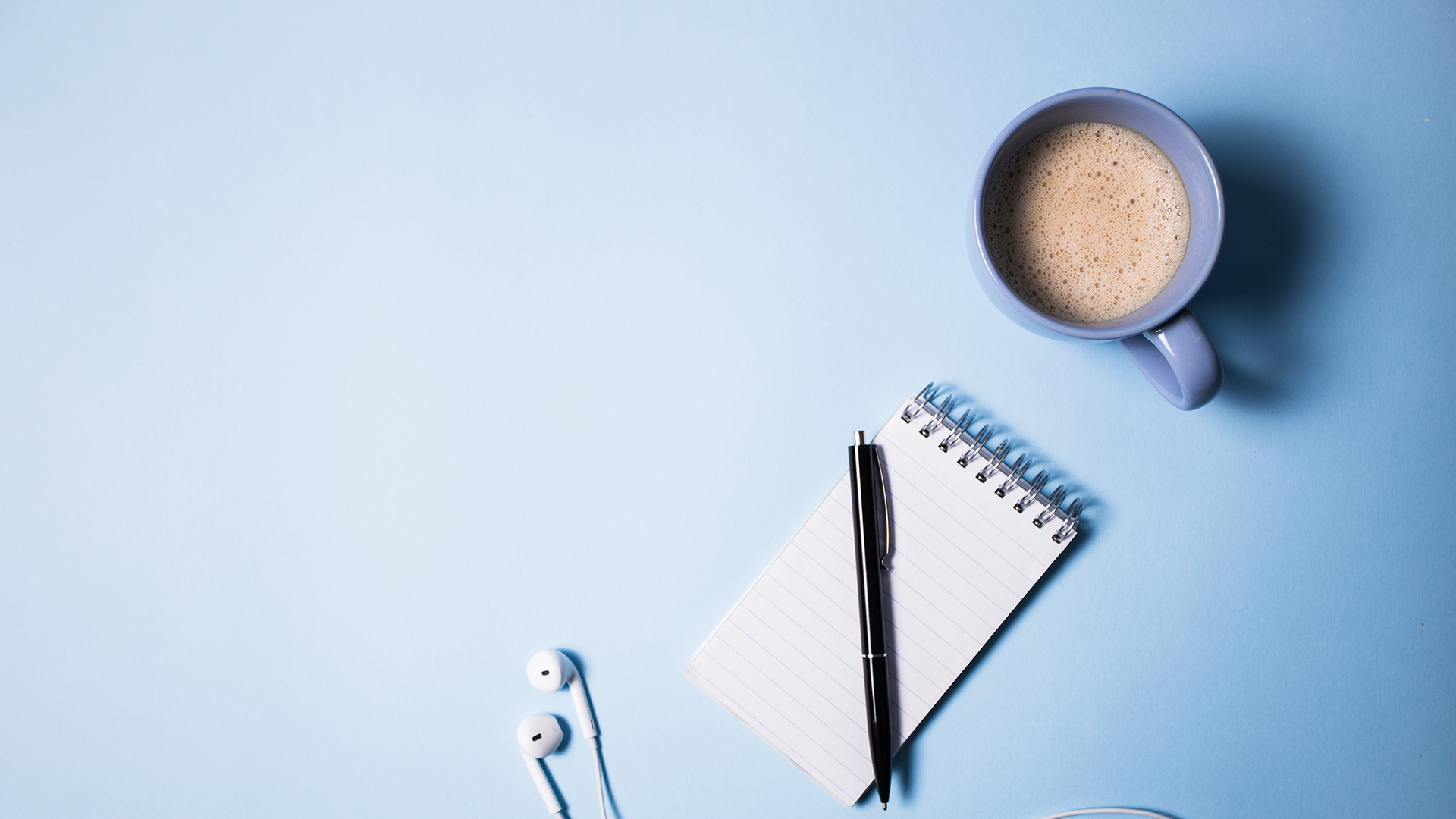 A blue mug of coffee, a notepad with a pen, and white earphones are arranged on a light blue surface. The items are positioned in the top right corner, leaving a large portion of the background empty.