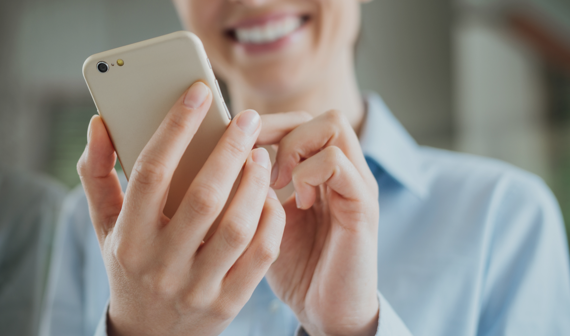 A person is smiling while using a smartphone. They are wearing a light blue shirt, and their focus is on the phone, which they hold with both hands. The background is blurred.