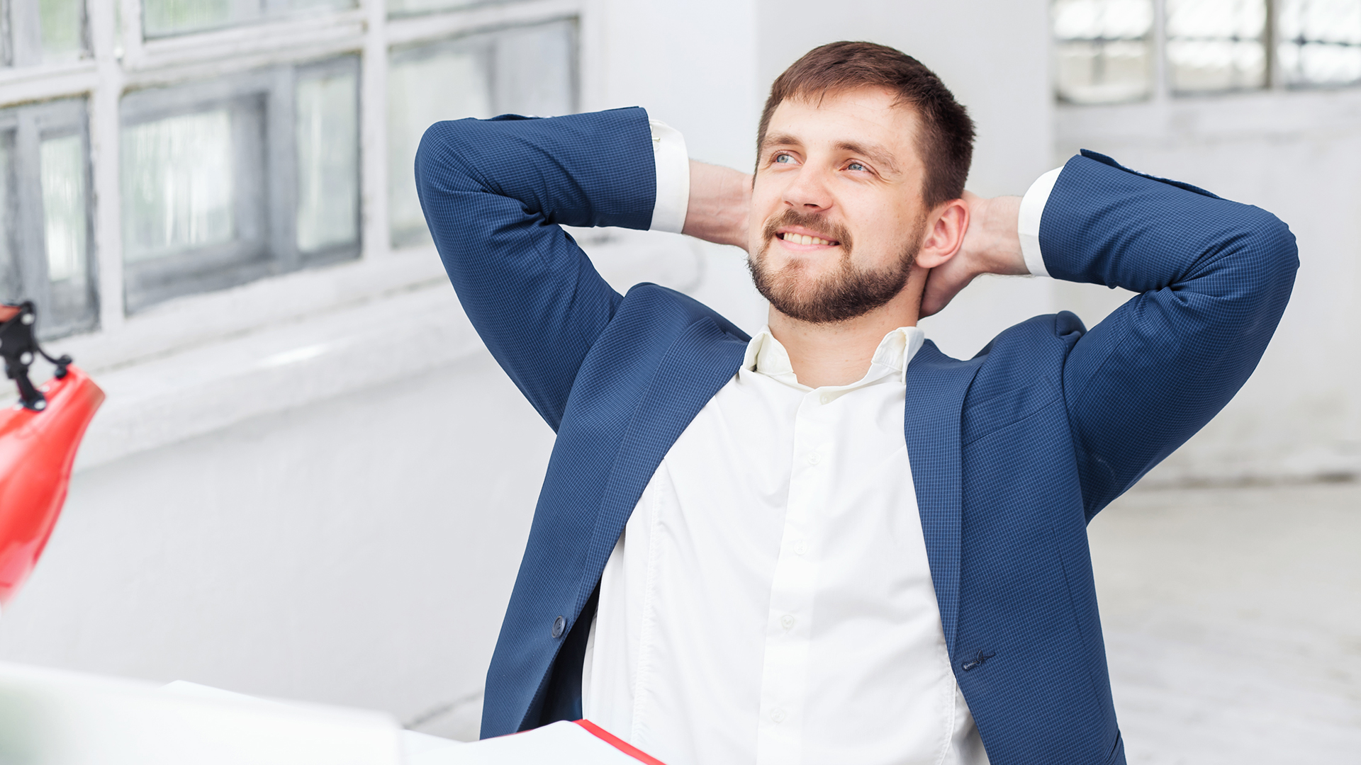 A man in a blue suit leans back in his chair with hands behind his head, smiling confidently.