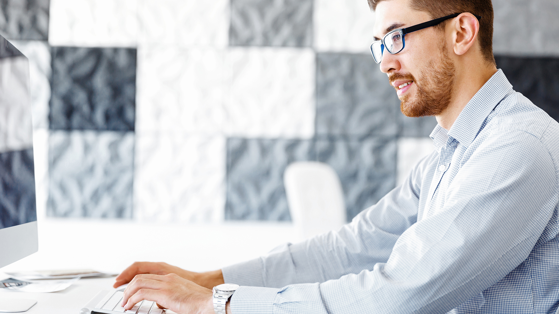 A man with glasses and a beard is sitting at a desk, typing on a computer keyboard. He is wearing a light-colored, long-sleeve shirt. The background features a pattern of gray and white squares.