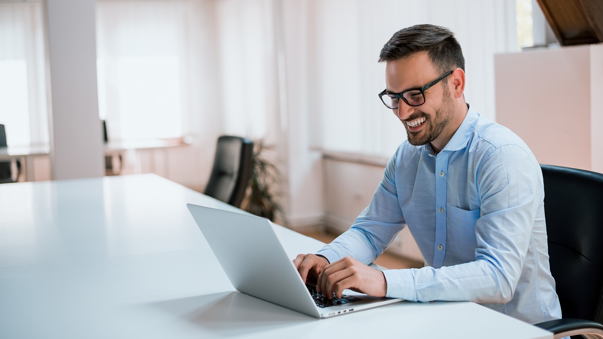 A man with short dark hair and glasses is sitting at a white table in a bright office, smiling while typing on a laptop. He is wearing a light blue shirt, and there are empty chairs and large windows in the background.