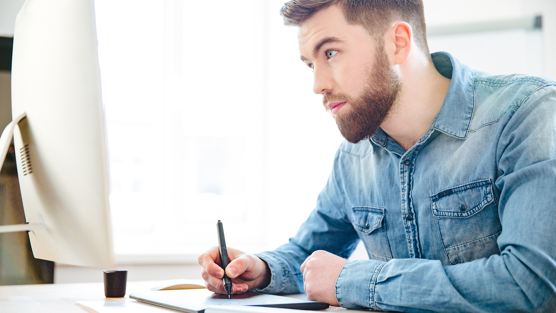 A man with a beard is sitting at a desk, attentively using a stylus on a graphic tablet. He is wearing a denim shirt and looking at a computer monitor. The background is bright, suggesting a well-lit room.