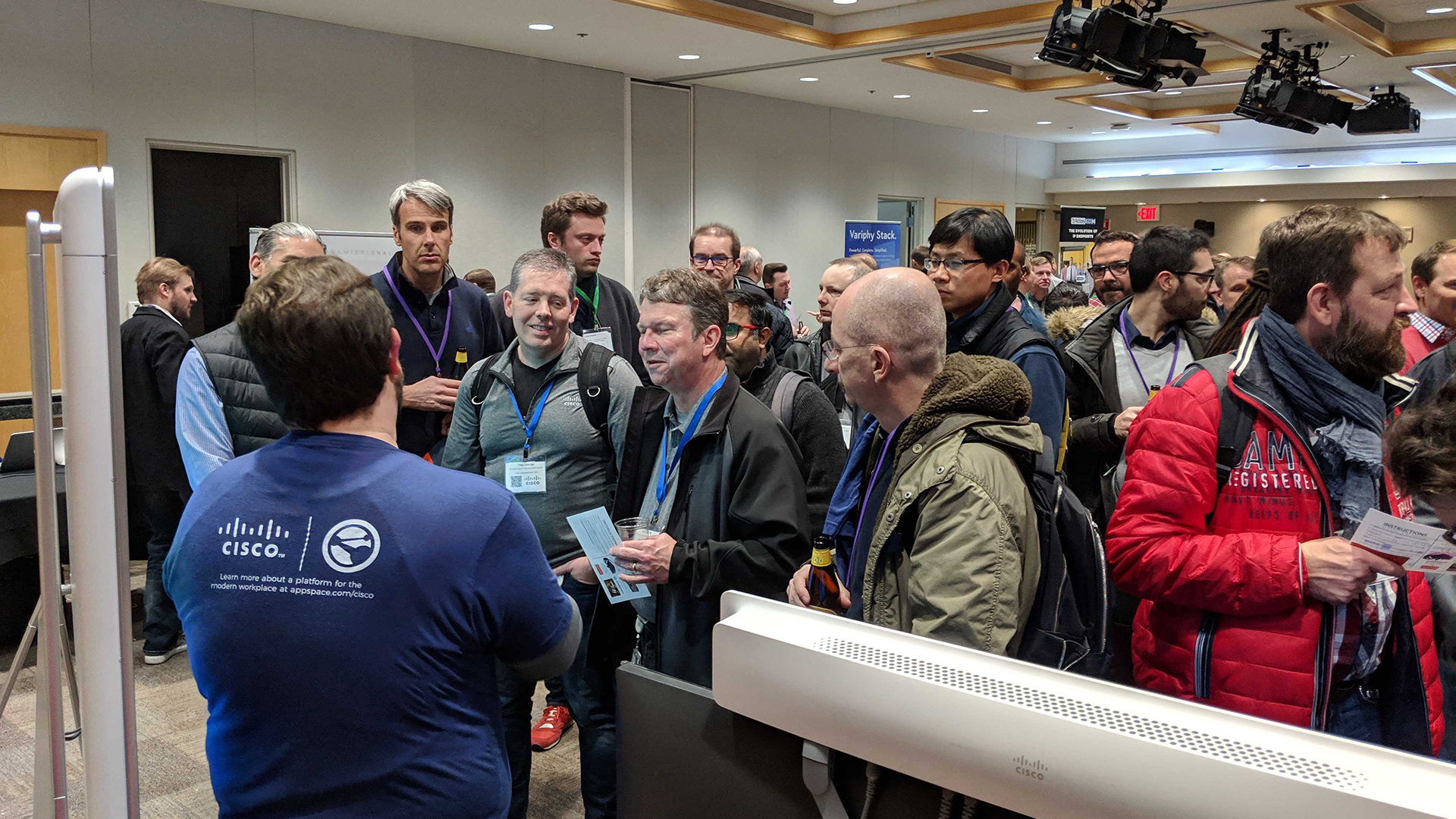 A bustling conference setting features attendees engaged in conversation. Some hold papers and wear ID badges. A person in the foreground sports a blue shirt with white logos. The venue is brightly lit with ceiling lights and signs on the walls.