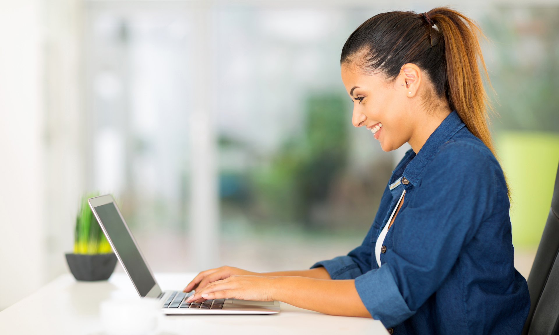 A person with long hair tied back is smiling while typing on a laptop. They are sitting at a desk with a small potted plant to the side. The background appears to be a bright, blurred indoor setting.