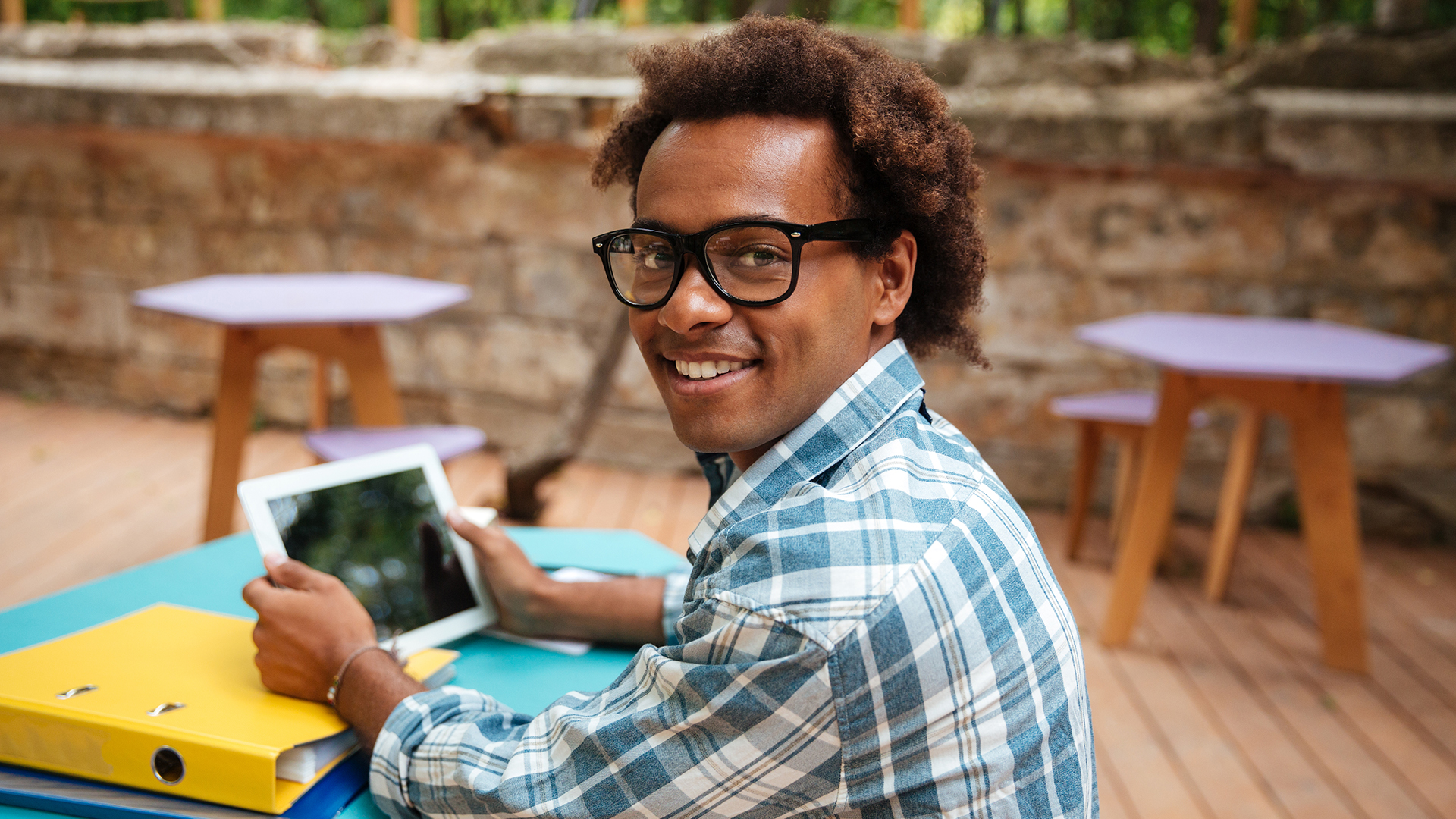 A person wearing glasses and a plaid shirt sits outside, smiling while holding a tablet. Nearby are folders on the table, and the background features a stone wall and wooden benches.