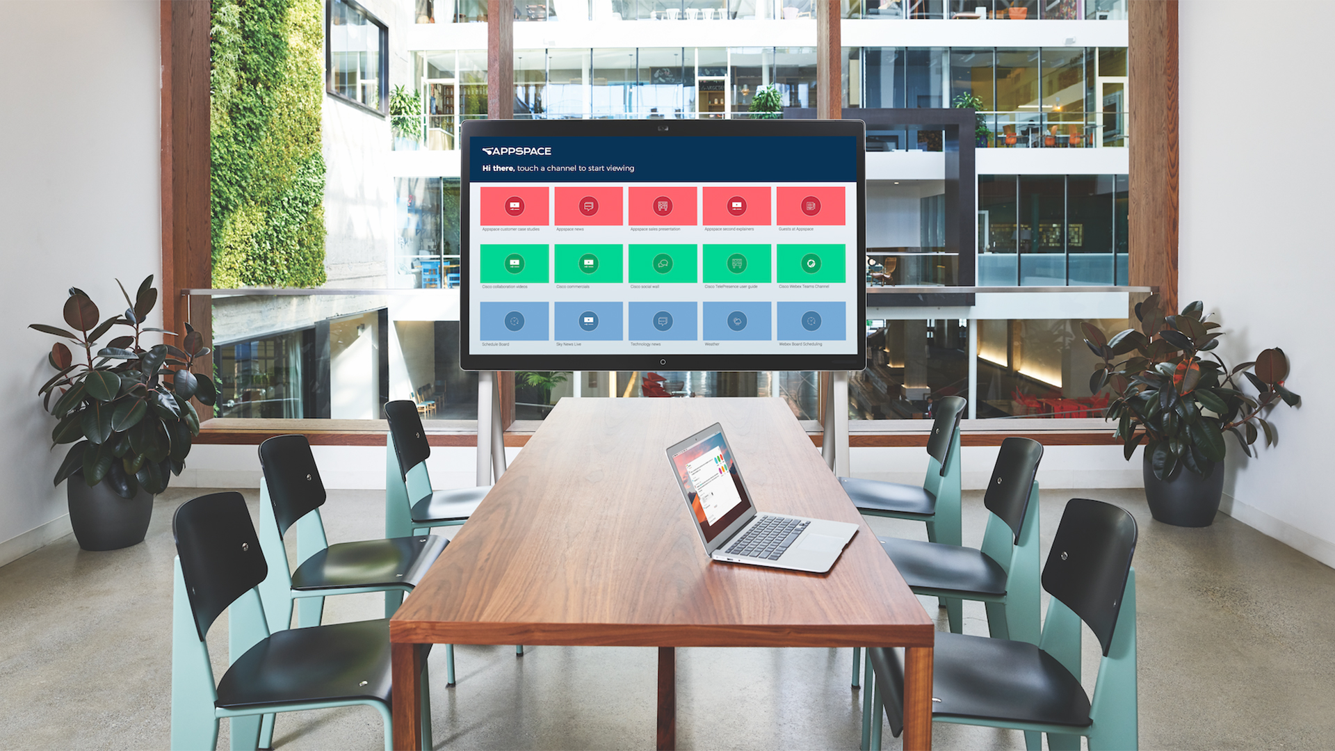 A modern meeting room with a wooden table, surrounded by black chairs. A laptop sits on the table, facing a large digital screen showing a grid interface with red, green, and blue panels. Large windows reveal an office buildings interior.