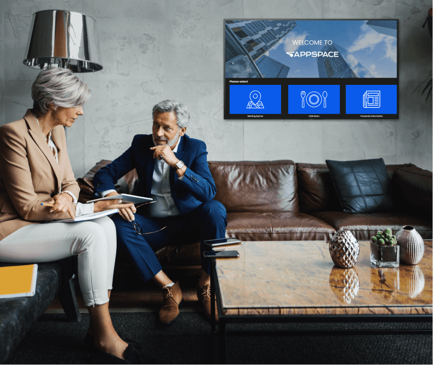 Two professionals in an office discussing work with a digital sign on the wall.