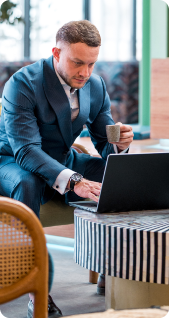 Professional man in suit drinking an espresso while working on his laptop in an open space.