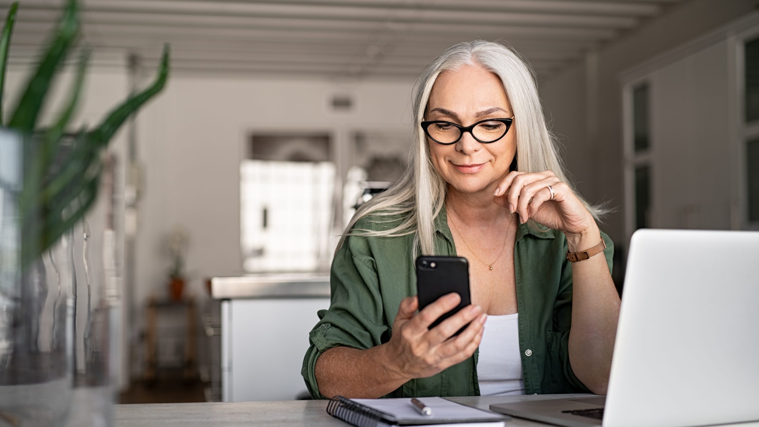 Happy senior woman using mobile employee app while working at home with laptop. Smiling cool old woman with white hair wearing eyeglasses sitting on chair at table and messaging with smartphone. Beautiful stylish elderly lady browsing site on cellphone.