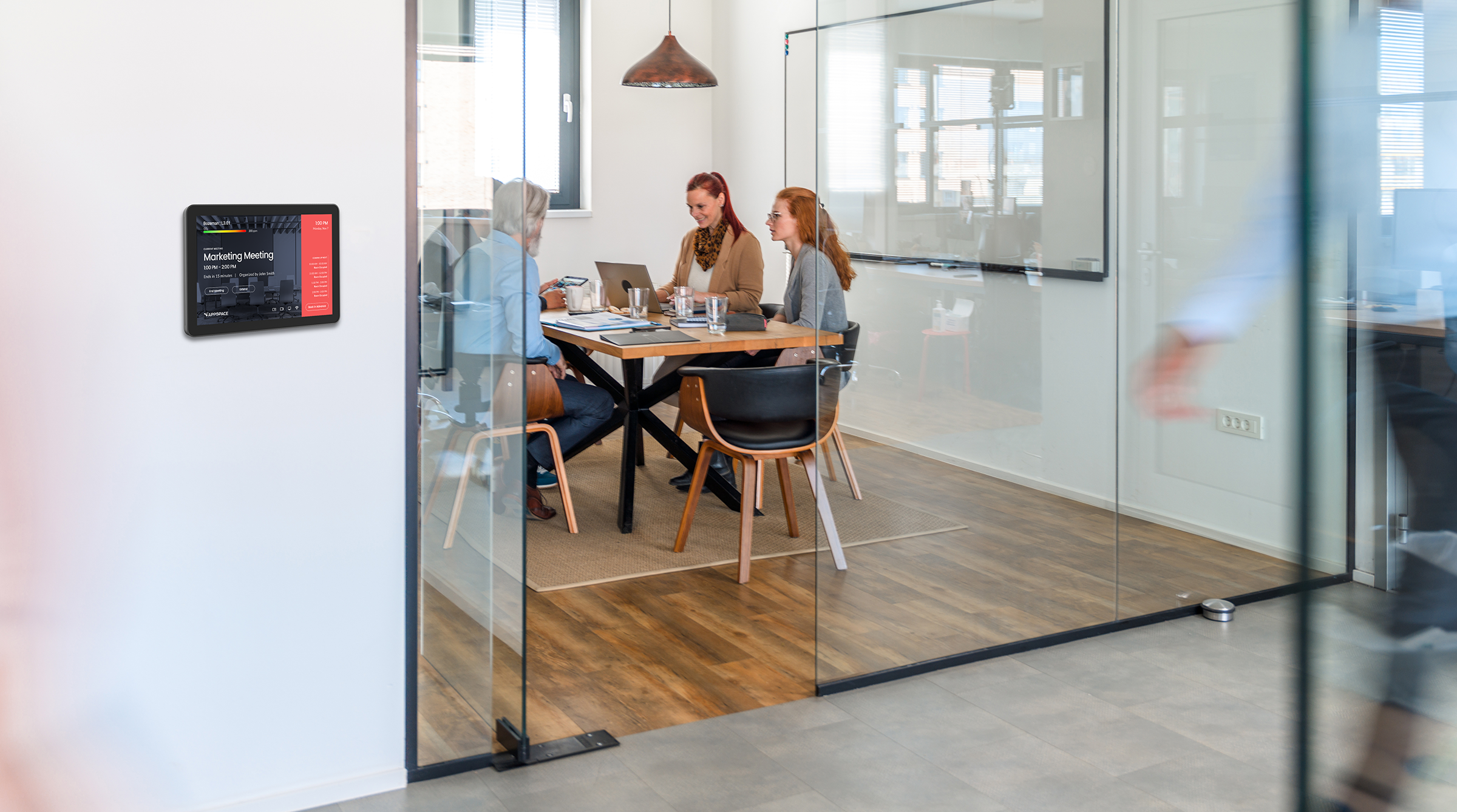Three people are seated around a table in a modern office meeting room with glass walls. A digital sign outside the room shows Room Occupied. Another person is visible walking outside the room. The space is lightly furnished with a hanging lamp.