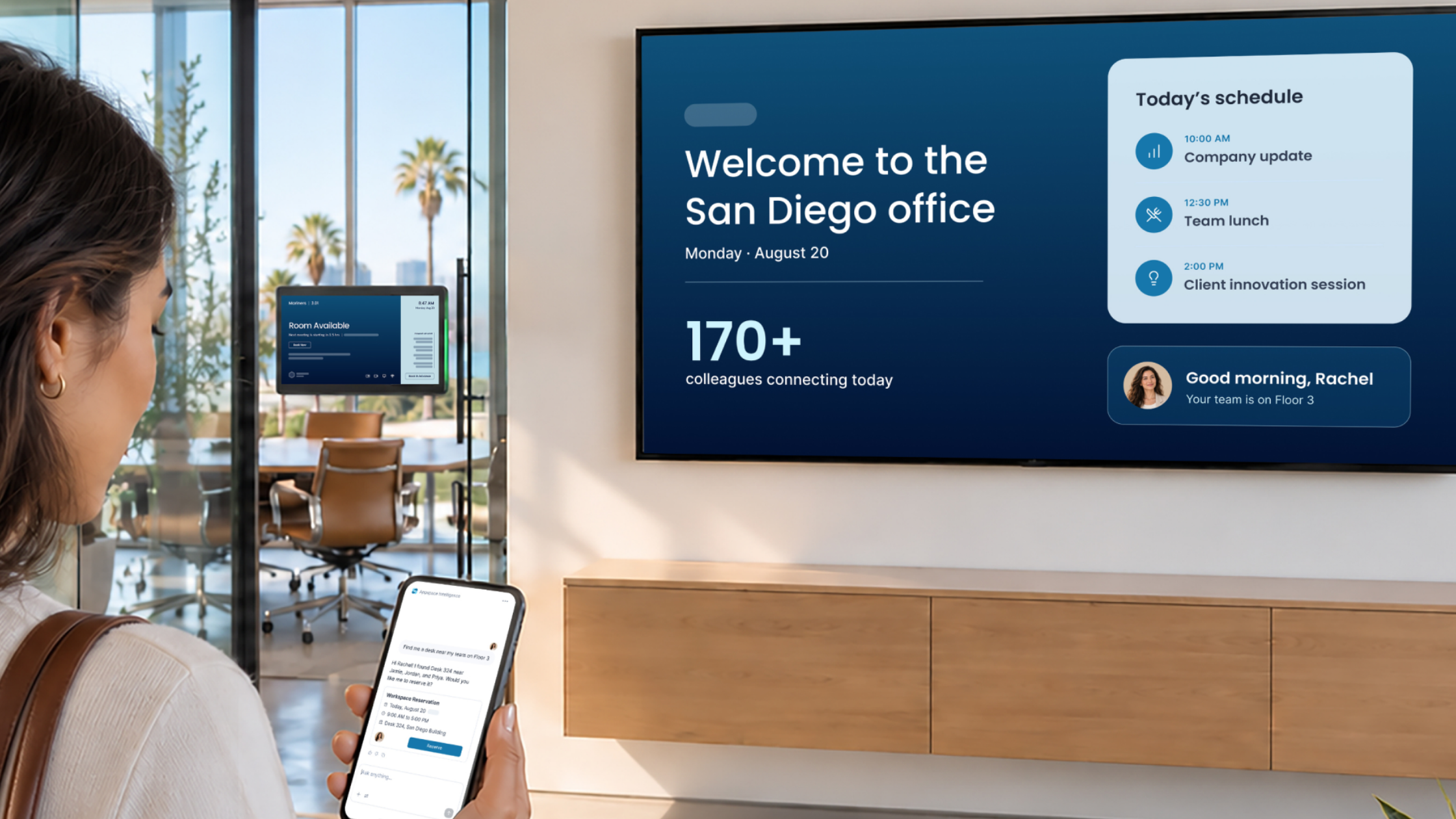 A woman stands in front of a digital sign in a workplace - it's using situational signage to display a personal welcome and today's agenda, which is also shown on the mobile phone she's holding.